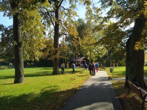 Widok na park, wśród drzew grupa seniorów rzuca lotkami do tarczy. W tle piknikowa altana